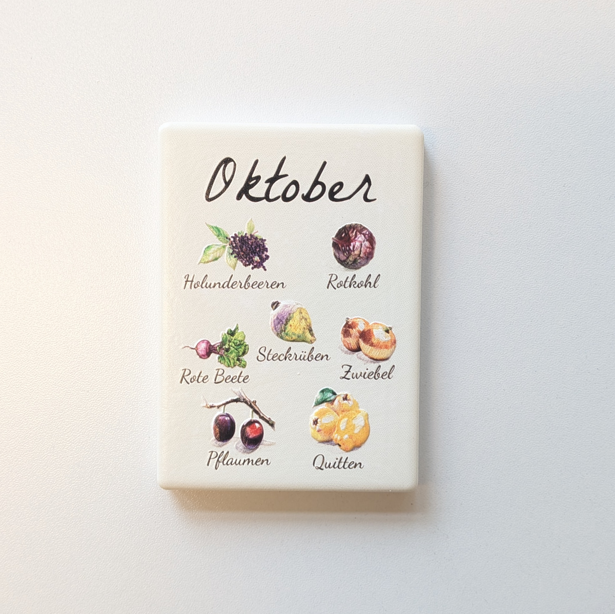 Saisonkalender Kühlschrankmagneten aus Keramik. Jeder Magnet repräsentiert einen Monat von Januar bis Dezember mit deutscher Beschriftung in geschwungener Schrift. Auf der cremefarbenen Oberfläche sind detailreiche, farbige Illustrationen von saisonalem Obst und Gemüse mit den jeweiligen Namen abgebildet.