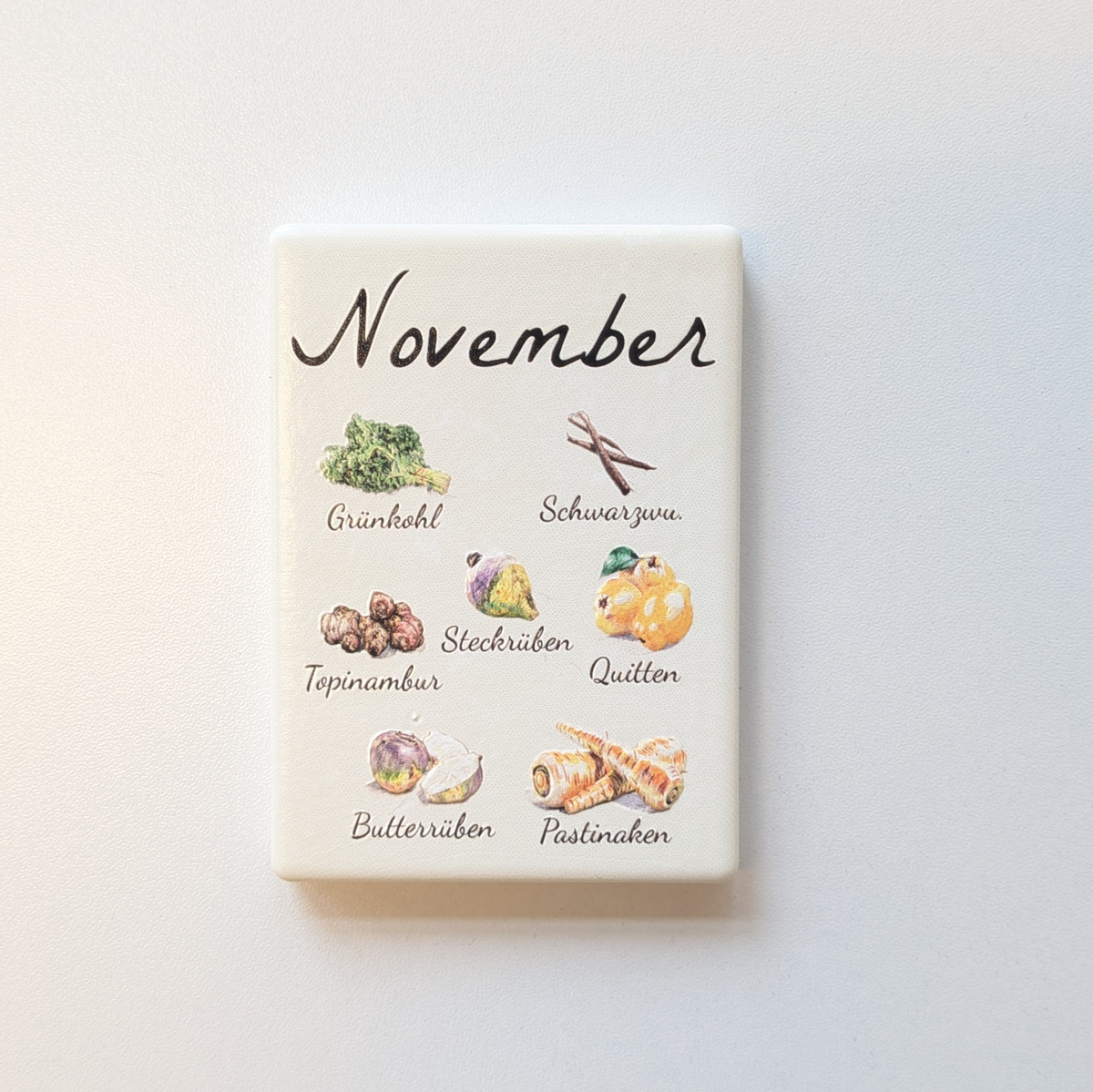 Saisonkalender Kühlschrankmagneten aus Keramik. Jeder Magnet repräsentiert einen Monat von Januar bis Dezember mit deutscher Beschriftung in geschwungener Schrift. Auf der cremefarbenen Oberfläche sind detailreiche, farbige Illustrationen von saisonalem Obst und Gemüse mit den jeweiligen Namen abgebildet.