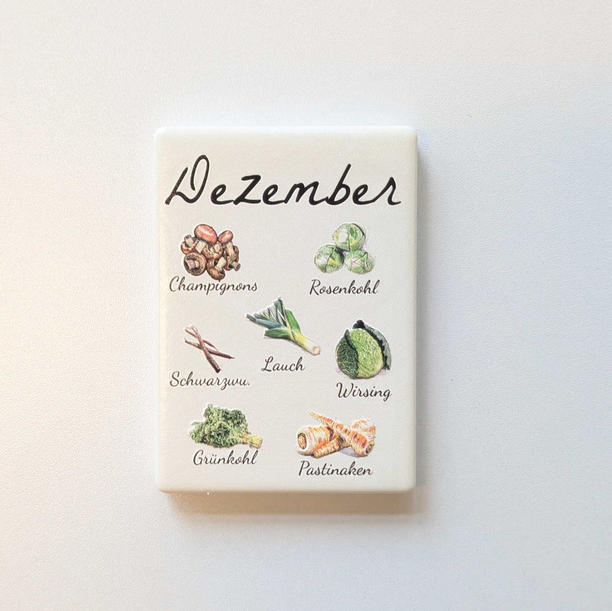 Saisonkalender Kühlschrankmagneten aus Keramik. Jeder Magnet repräsentiert einen Monat von Januar bis Dezember mit deutscher Beschriftung in geschwungener Schrift. Auf der cremefarbenen Oberfläche sind detailreiche, farbige Illustrationen von saisonalem Obst und Gemüse mit den jeweiligen Namen abgebildet.