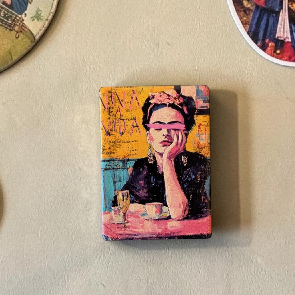 Künstlerisches Porträt einer Frau mit Blumen im Haar und einem pinkfarbenen Pinselstrich über den Augen. Im Hintergrund steht der Schriftzug „VIVA LA VIDA“ auf gelbem, strukturiertem Grund.