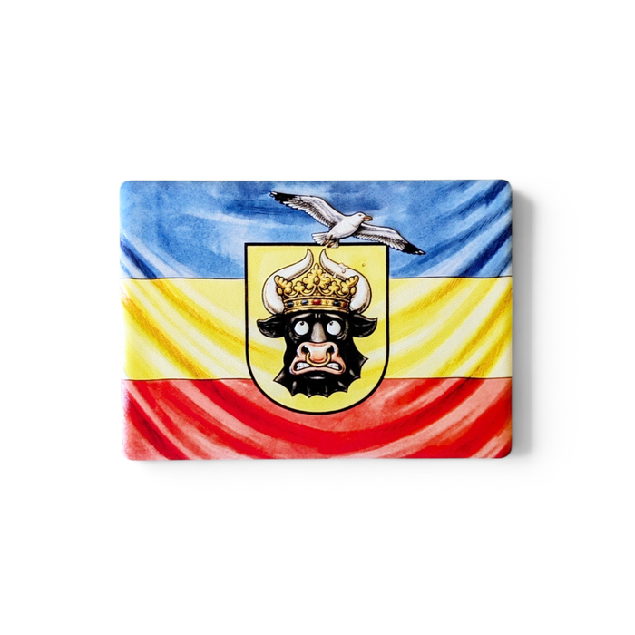 Rechteckiger Keramik-Kühlschrankmagnet (7x5 cm) im Querformat mit seidenmatter Glasur. Abgebildet ist die mecklenburgische Flagge in Blau, Gelb und Rot mit einem lustig dreinschauenden Stierkopf mit goldener Krone, auf dem eine Möwe sitzt, im spürbaren 3D-Relief.