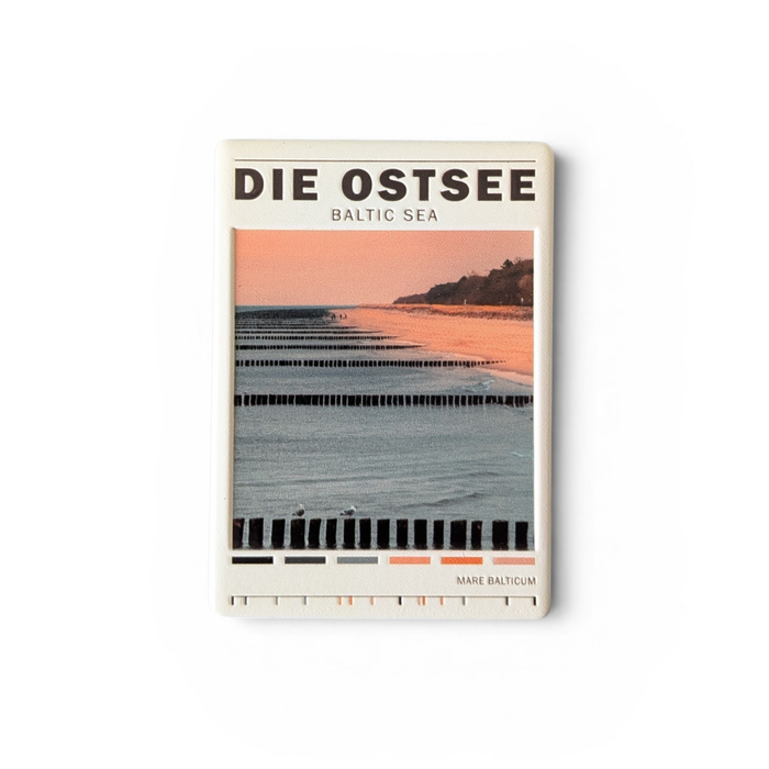 Rechteckiger Keramik-Kühlschrankmagnet (7x5 cm) mit einem Ostsee-Strandmotiv bei weichem Licht, hölzernen Buhnen im Wasser, dem Schriftzug 'DIE OSTSEE BALTIC SEA' und einer kleinen Farbpalette am unteren Rand mit spürbarem 3D-Relief.