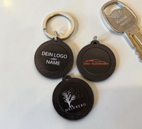 Hand hält einen runden, schwarzen NFC-Schlüsselanhänger aus Holz mit der
Aufschrift "DEIN LOGO -oder- NAME". Im Hintergrund eine kleine grüne Pflanze
auf hellem Tisch.