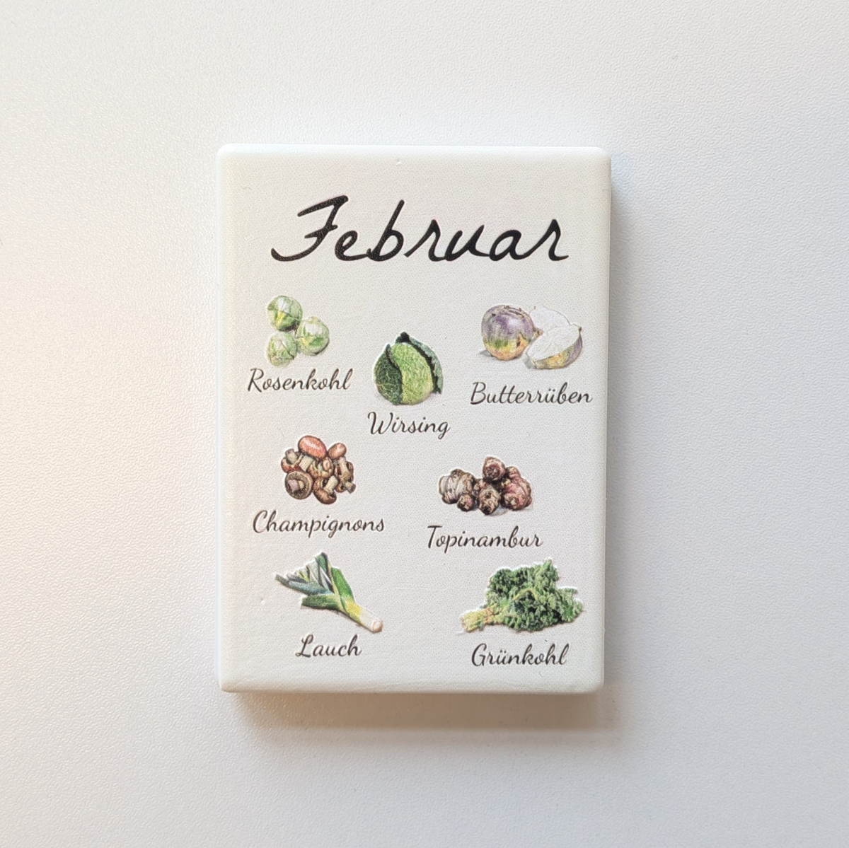 Saisonkalender Kühlschrankmagneten aus Keramik. Jeder Magnet repräsentiert einen Monat von Januar bis Dezember mit deutscher Beschriftung in geschwungener Schrift. Auf der cremefarbenen Oberfläche sind detailreiche, farbige Illustrationen von saisonalem Obst und Gemüse mit den jeweiligen Namen abgebildet.