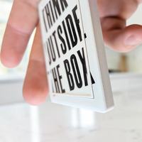 "Rechteckiger Keramik-Kühlschrankmagnet (7x5 cm) in Schwarz-Weiß mit seidenmatter Glasur. Gezeigt wird der Schriftzug 'THINK OUTSIDE THE BOX', dessen Buchstaben im fühlbaren 3D-Relief über einen erhabenen, dünnen Rahmen hinausragen."