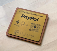 Quadratischer PayPal-Tischaufsteller aus dunklem Holz mit einer gravierten, gold gebürsteten Kunststoffplatte. Auf der Platte sind ein QR-Code, ein NFC-Symbol und das PayPal-Logo zu sehen, perfekt geeignet für kontaktlose Zahlungen auf einer Ladentheke.