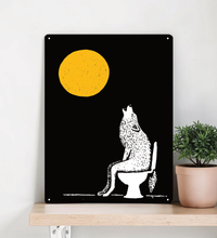 Ein hochformatiges, schwarzes Metallschild steht dekorativ auf einem hellen Holzregal vor einer beigen Wand. Das Motiv zeigt einen großen, strukturierten gelben Vollmond und die weiße Silhouette eines Wolfs, der auf einer Toilette sitzt und nach oben heult. 