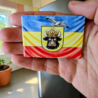 Rechteckiger Keramik-Kühlschrankmagnet (7x5 cm) im Querformat mit seidenmatter Glasur. Abgebildet ist die mecklenburgische Flagge in Blau, Gelb und Rot mit einem lustig dreinschauenden Stierkopf mit goldener Krone, auf dem eine Möwe sitzt, im spürbaren 3D-Relief.