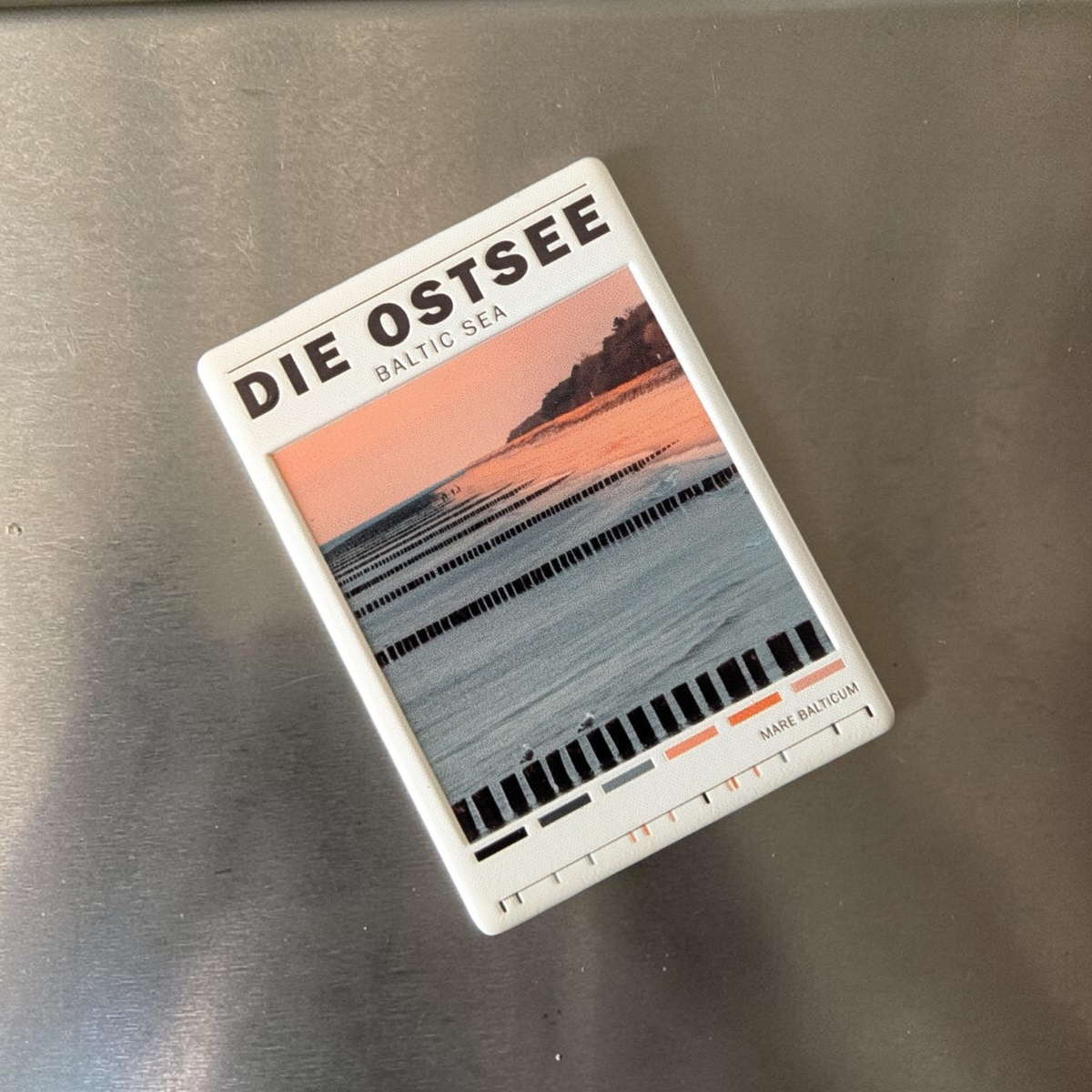 Rechteckiger Keramik-Kühlschrankmagnet (7x5 cm) mit einem Ostsee-Strandmotiv bei weichem Licht, hölzernen Buhnen im Wasser, dem Schriftzug 'DIE OSTSEE BALTIC SEA' und einer kleinen Farbpalette am unteren Rand mit spürbarem 3D-Relief.