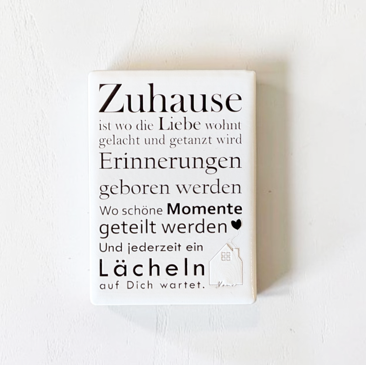 Rechteckiger Keramik-Kühlschrankmagnet (7x5 cm) mit seidenmatter Glasur. Auf hellem Grund steht der schwarze Spruch 'Zuhause ist wo die Liebe wohnt...' neben einer kleinen Haus-Illustration mit Herz im spürbaren 3D-Relief.