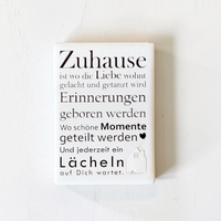 Rechteckiger Keramik-Kühlschrankmagnet (7x5 cm) mit seidenmatter Glasur. Auf hellem Grund steht der schwarze Spruch 'Zuhause ist wo die Liebe wohnt...' neben einer kleinen Haus-Illustration mit Herz im spürbaren 3D-Relief.