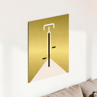 Minimalistische Bauhaus-Illustration eines Fahrrads in der Vorderansicht auf einer gebürsteten, gold-glänzenden Aluminiumplatte (Alu-Dibond). Das Design zeigt klare weiße und schwarze Linien vor einem edlen Metallhintergrund. Das Bild lehnt modern an einer weißen Wand zwischen zwei Zimmerpflanzen.
