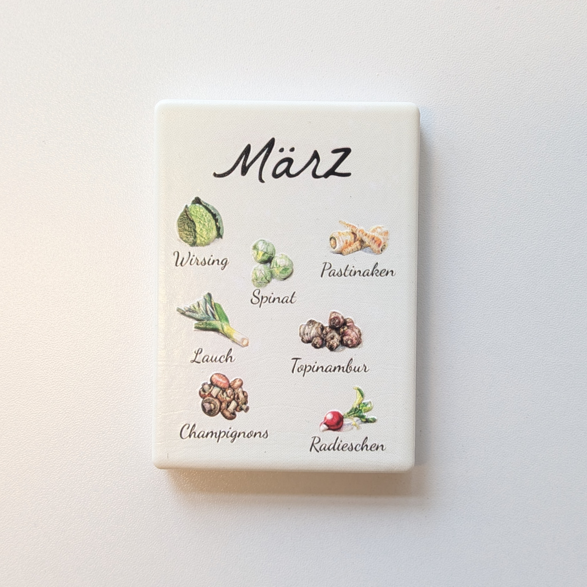 Saisonkalender Kühlschrankmagneten aus Keramik. Jeder Magnet repräsentiert einen Monat von Januar bis Dezember mit deutscher Beschriftung in geschwungener Schrift. Auf der cremefarbenen Oberfläche sind detailreiche, farbige Illustrationen von saisonalem Obst und Gemüse mit den jeweiligen Namen abgebildet.