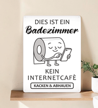 Ein rechteckiges, weißes Metallschild im Hochformat mit dem schwarzen Text „Das hier ist kein Internetcafé! Beeilt euch, macht euer Geschäft und geht wieder!“. Das Schild steht aufrecht vor einem schlichten, hellen Hintergrund.