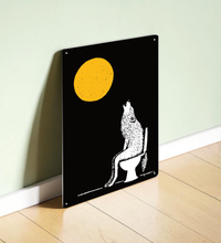 Ein hochformatiges, schwarzes Metallschild steht dekorativ auf einem hellen Holzregal vor einer beigen Wand. Das Motiv zeigt einen großen, strukturierten gelben Vollmond und die weiße Silhouette eines Wolfs, der auf einer Toilette sitzt und nach oben heult. 