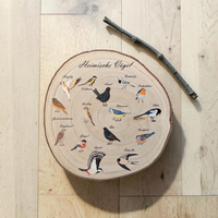 Eine runde Holzscheibe aus hellem Paulownia-Holz mit echter Baumrinde. Auf der Oberfläche sind im Kreis 14 verschiedene heimische Vogelarten wie Blaumeise, Rotkehlchen und Specht mit ihren Namen farbig aufgedruckt. Oben steht der geschwungene Schriftzug "Heimische Vögel". Der Druck hat ein fühlbares 0,3 mm Relief.
