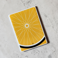 Rechteckiger Keramik-Kühlschrankmagnet (7x5 cm) in warmem Gelb. Gezeigt wird ein minimalistisches Fahrradrad mit feinen weißen Speichen, einem schwarzen Reifen am unteren Rand und einer winzigen menschlichen Silhouette im spürbaren 3D-Relief.