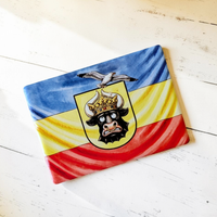 Rechteckiger Keramik-Kühlschrankmagnet (7x5 cm) im Querformat mit seidenmatter Glasur. Abgebildet ist die mecklenburgische Flagge in Blau, Gelb und Rot mit einem lustig dreinschauenden Stierkopf mit goldener Krone, auf dem eine Möwe sitzt, im spürbaren 3D-Relief.