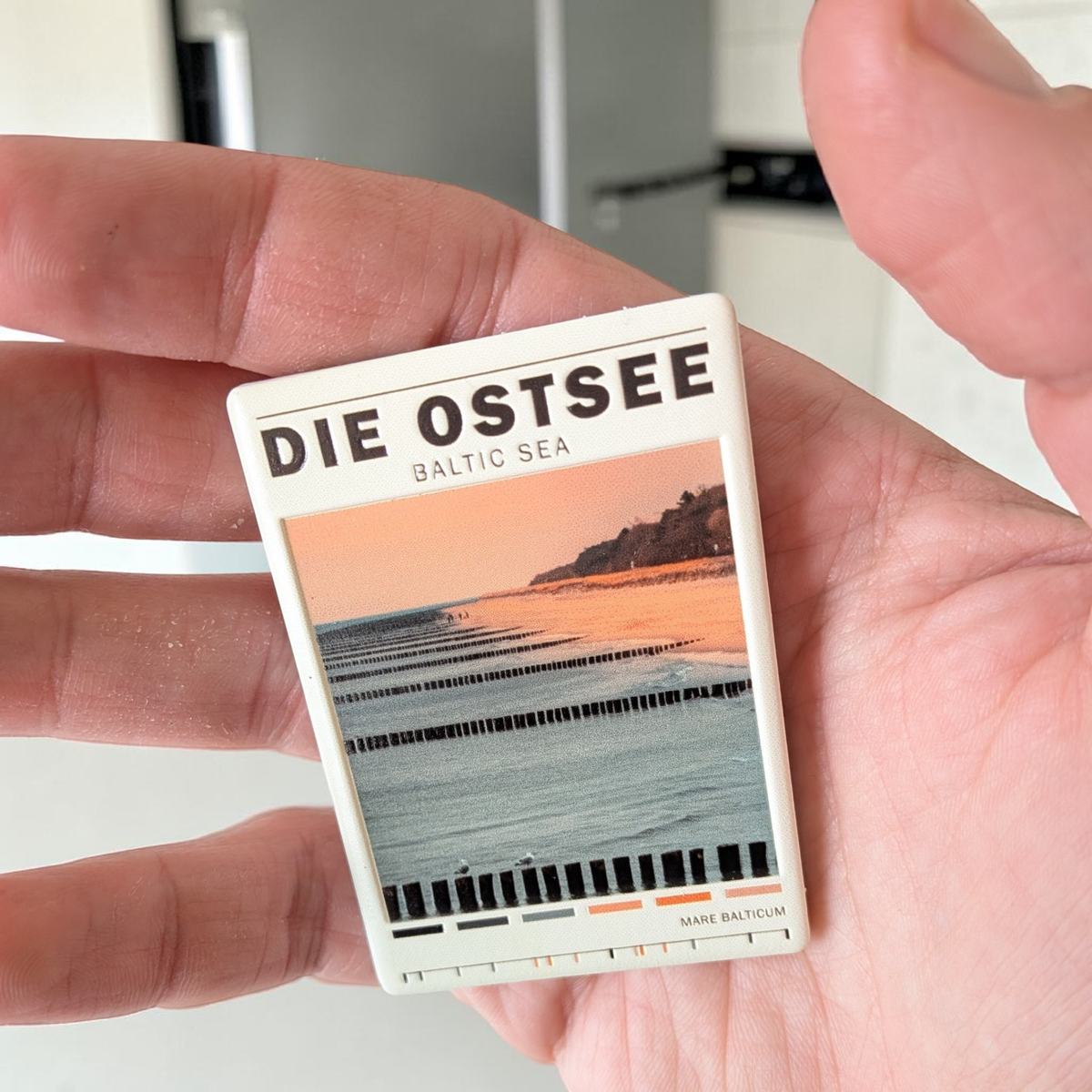 Rechteckiger Keramik-Kühlschrankmagnet (7x5 cm) mit einem Ostsee-Strandmotiv bei weichem Licht, hölzernen Buhnen im Wasser, dem Schriftzug 'DIE OSTSEE BALTIC SEA' und einer kleinen Farbpalette am unteren Rand mit spürbarem 3D-Relief.