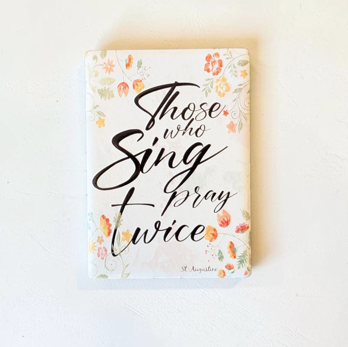 Rechteckiger Keramik-Kühlschrankmagnet (7x5 cm) mit seidenmatter Glasur. Auf hellem Grund mit zarten orange-gelben Aquarell-Blumen steht der schwarze Handlettering-Schriftzug 'Who sings prays twice' im spürbaren 3D-Relief