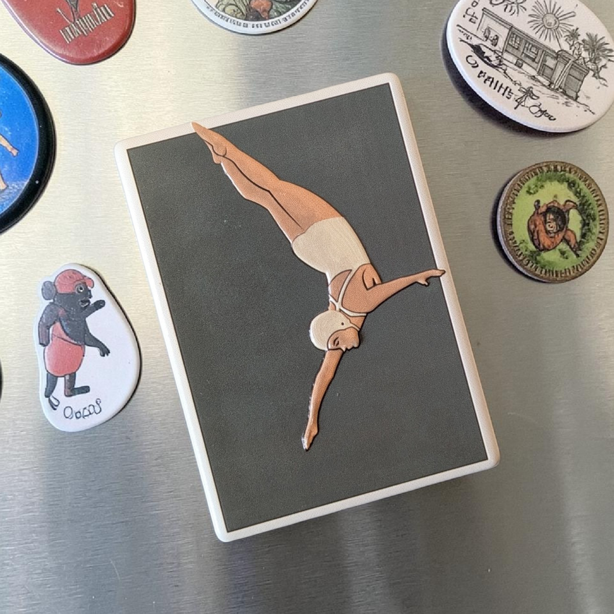 Rechteckiger weißer Keramikmagnet mit einer minimalistischen Illustration einer Turmspringerin im Sprung. Das Motiv hat ein fühlbares Relief und eine edle, seidenmatte Oberfläche.