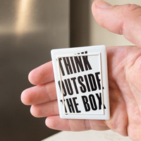 "Rechteckiger Keramik-Kühlschrankmagnet (7x5 cm) in Schwarz-Weiß mit seidenmatter Glasur. Gezeigt wird der Schriftzug 'THINK OUTSIDE THE BOX', dessen Buchstaben im fühlbaren 3D-Relief über einen erhabenen, dünnen Rahmen hinausragen."