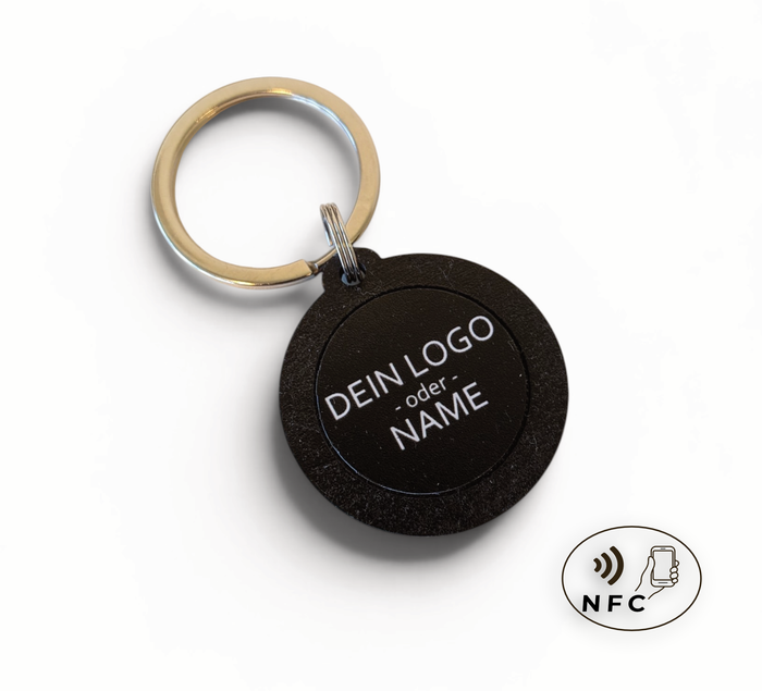 Hand hält einen runden, schwarzen NFC-Schlüsselanhänger aus Holz mit der
Aufschrift "DEIN LOGO -oder- NAME". Im Hintergrund eine kleine grüne Pflanze
auf hellem Tisch.