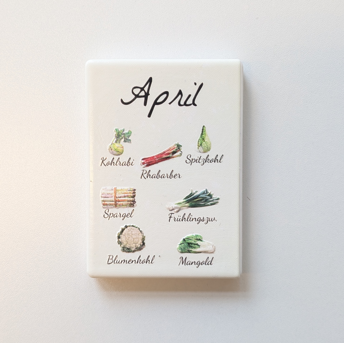 Saisonkalender Kühlschrankmagneten aus Keramik. Jeder Magnet repräsentiert einen Monat von Januar bis Dezember mit deutscher Beschriftung in geschwungener Schrift. Auf der cremefarbenen Oberfläche sind detailreiche, farbige Illustrationen von saisonalem Obst und Gemüse mit den jeweiligen Namen abgebildet.