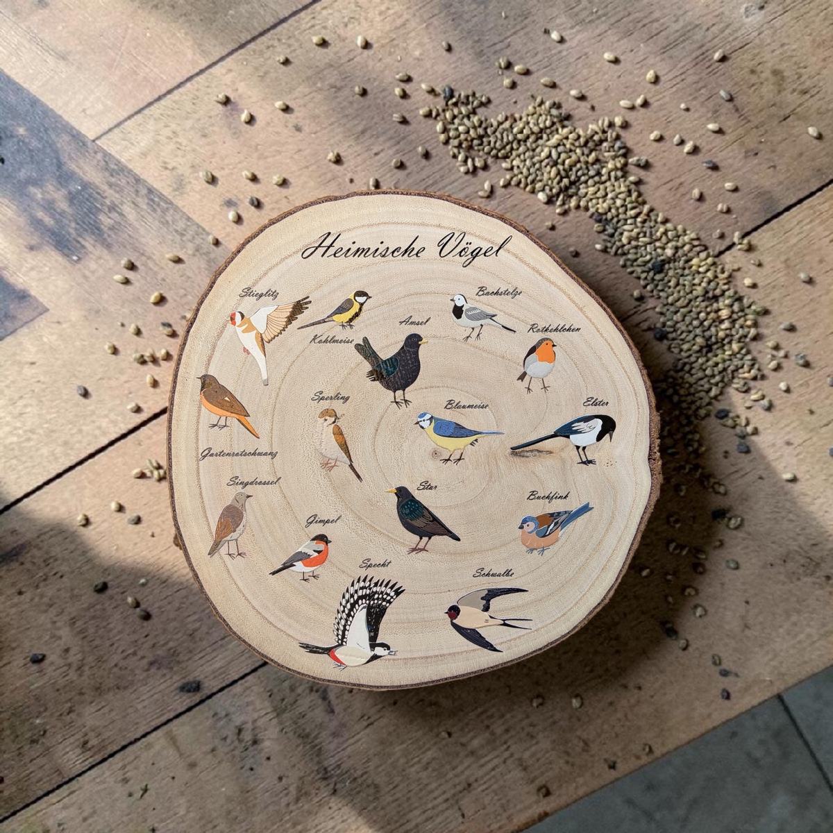 Eine runde Holzscheibe aus hellem Paulownia-Holz mit echter Baumrinde. Auf der Oberfläche sind im Kreis 14 verschiedene heimische Vogelarten wie Blaumeise, Rotkehlchen und Specht mit ihren Namen farbig aufgedruckt. Oben steht der geschwungene Schriftzug "Heimische Vögel". Der Druck hat ein fühlbares 0,3 mm Relief.