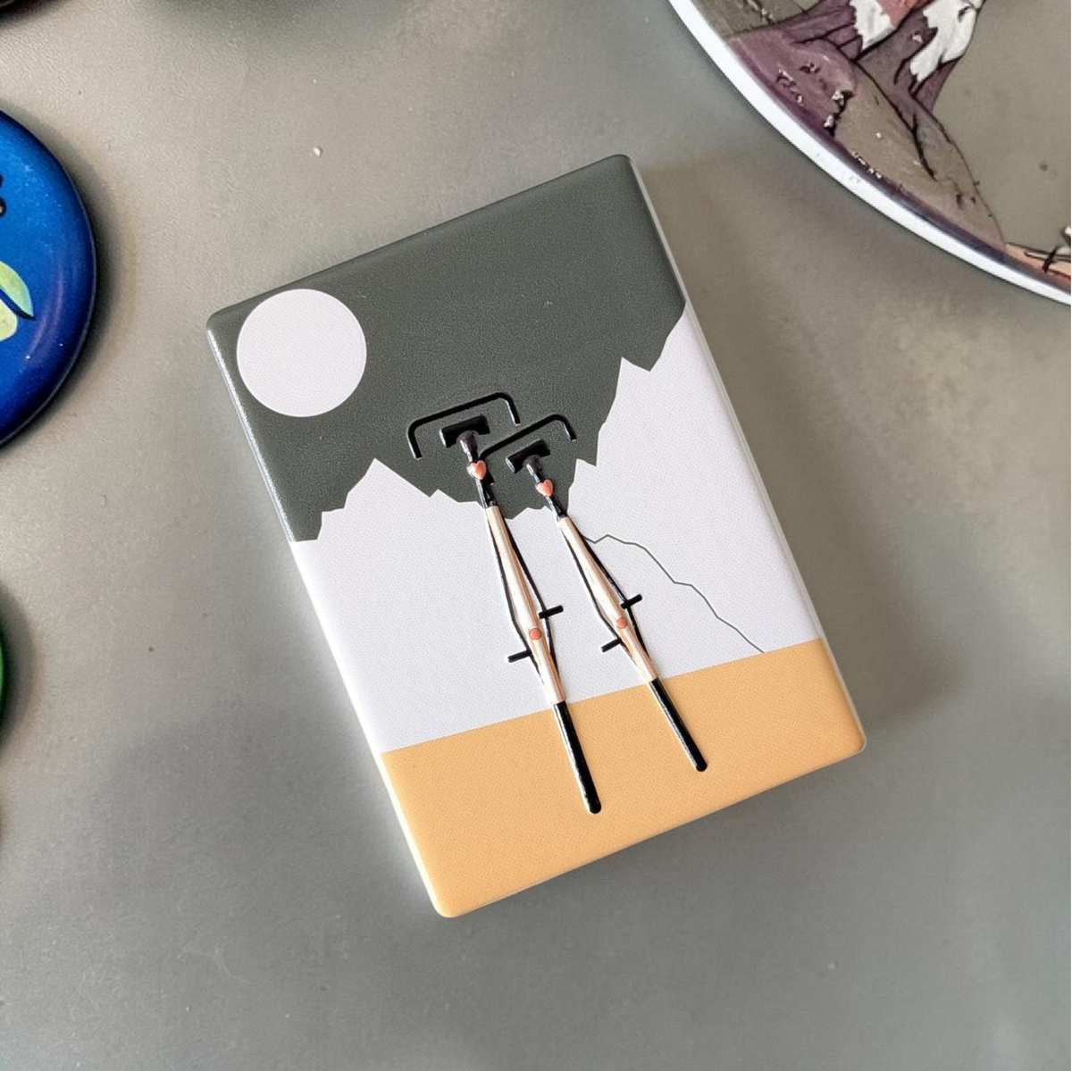Rechteckiger Keramik-Kühlschrankmagnet (7x5 cm) mit zwei schwarzen Fahrrädern im Vordergrund vor weißen Bergen, einem graugrünen Himmel mit weißem Vollmond und einem ockergelben Boden mit spürbarem 3D-Relief.