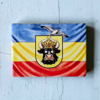 Rechteckiger Keramik-Kühlschrankmagnet (7x5 cm) im Querformat mit seidenmatter Glasur. Abgebildet ist die mecklenburgische Flagge in Blau, Gelb und Rot mit einem lustig dreinschauenden Stierkopf mit goldener Krone, auf dem eine Möwe sitzt, im spürbaren 3D-Relief.
