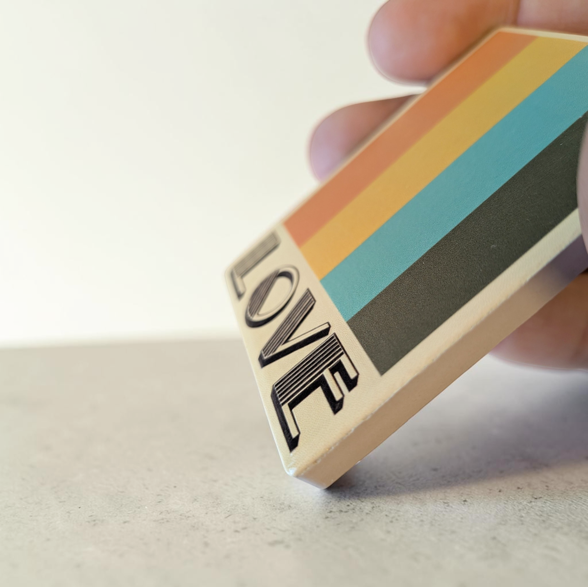 Rechteckiger Keramik-Kühlschrankmagnet (7x5 cm) mit seidenmatter Glasur. Auf beigem Grund verlaufen vier vertikale Streifen in Orange, Gelb, Petrol und Braun über dem schwarzen Schriftzug 'LOVE' im fühlbaren 3D-Relief.