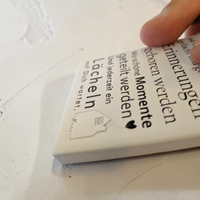 Rechteckiger Keramik-Kühlschrankmagnet (7x5 cm) mit seidenmatter Glasur. Auf hellem Grund steht der schwarze Spruch 'Zuhause ist wo die Liebe wohnt...' neben einer kleinen Haus-Illustration mit Herz im spürbaren 3D-Relief.