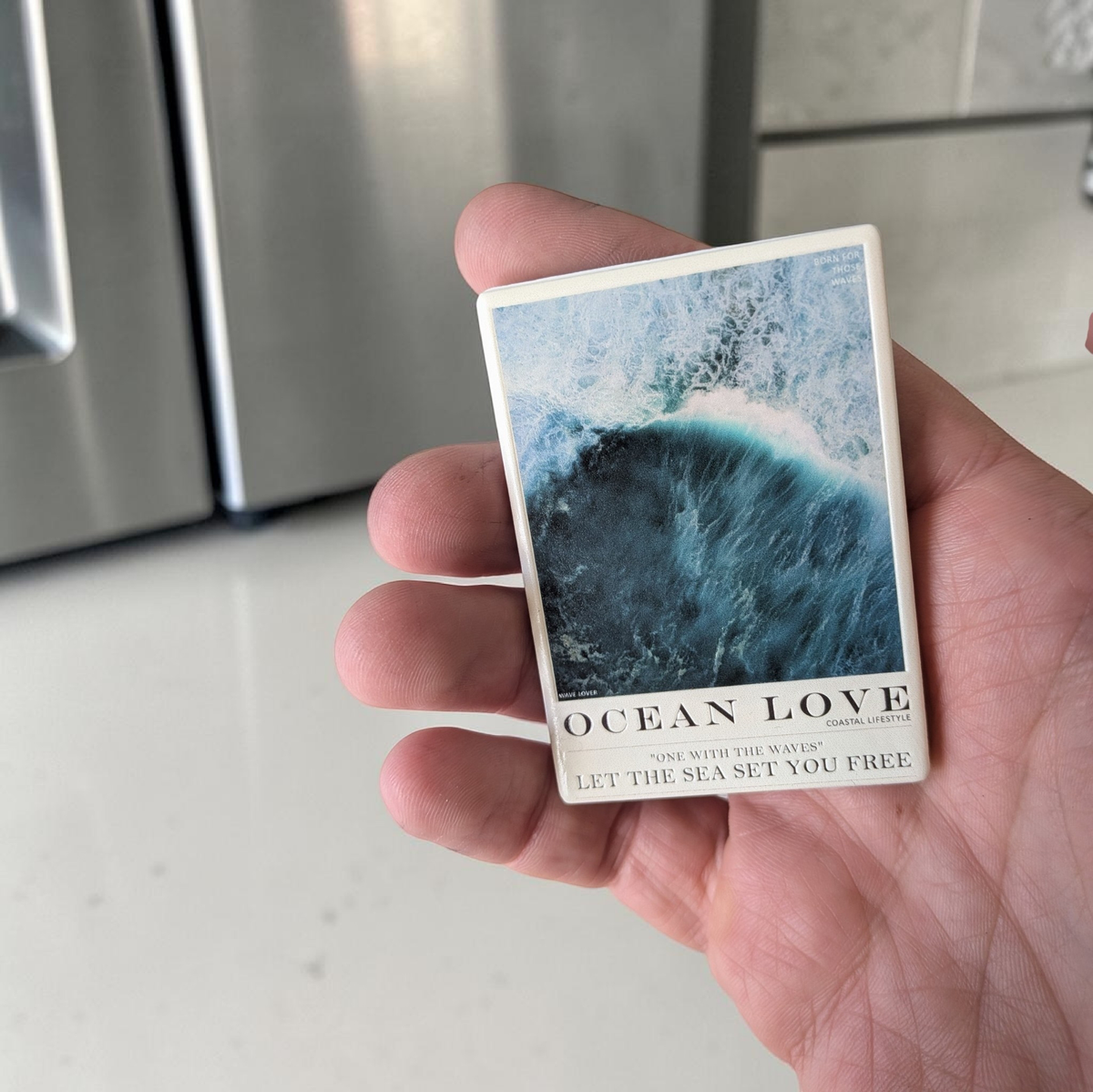 Rechteckiger Keramik-Kühlschrankmagnet (7x5 cm) mit seidenmatter Glasur. Abgebildet ist eine stilisierte Meereswelle in verschiedenen Blautönen mit dem Schriftzug 'OCEAN LOVE' im spürbaren 3D-Relief.