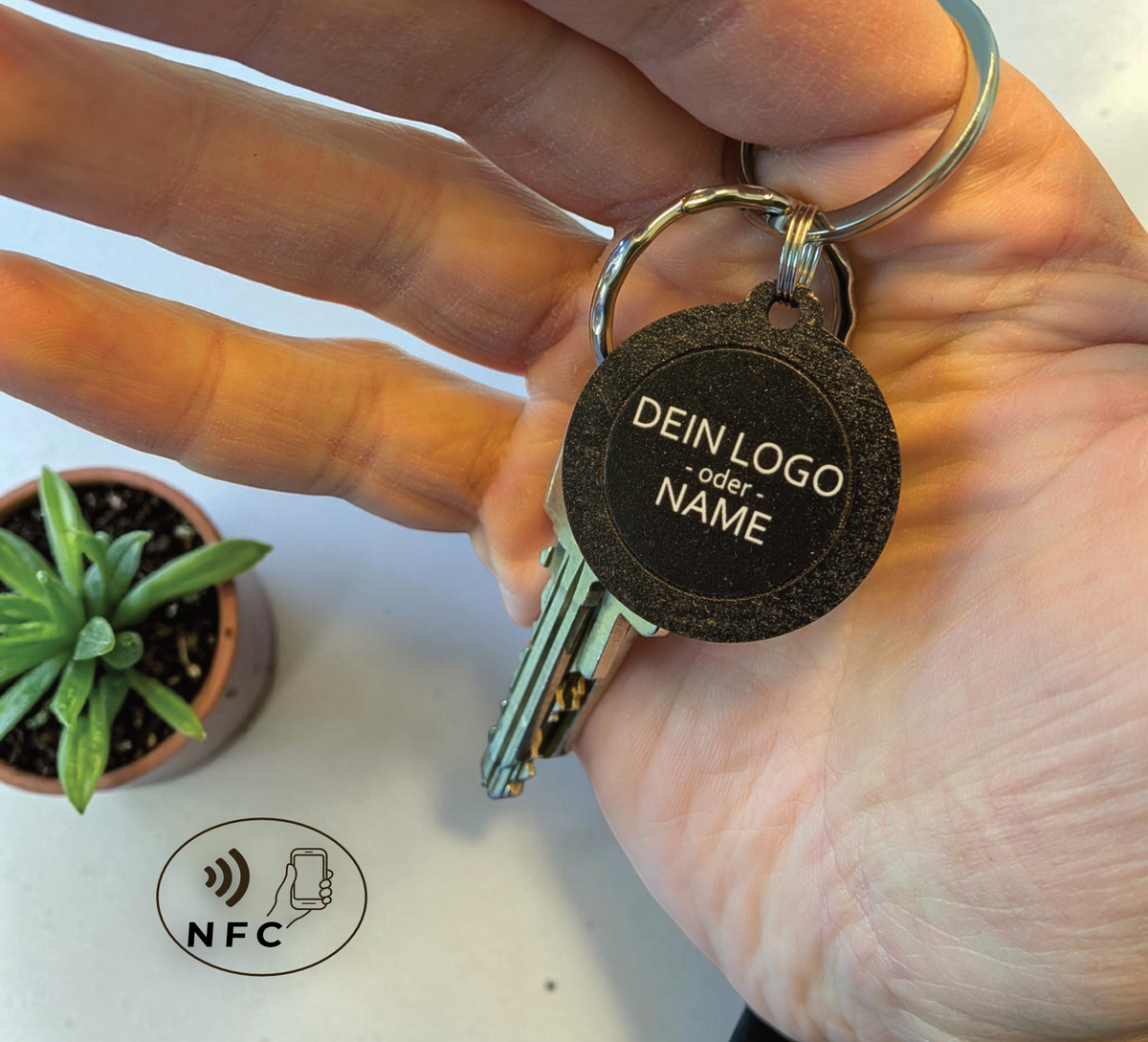 Hand hält einen runden, schwarzen NFC-Schlüsselanhänger aus Holz mit der
Aufschrift "DEIN LOGO -oder- NAME". Im Hintergrund eine kleine grüne Pflanze
auf hellem Tisch.