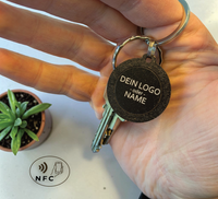 Hand hält einen runden, schwarzen NFC-Schlüsselanhänger aus Holz mit der
Aufschrift "DEIN LOGO -oder- NAME". Im Hintergrund eine kleine grüne Pflanze
auf hellem Tisch.