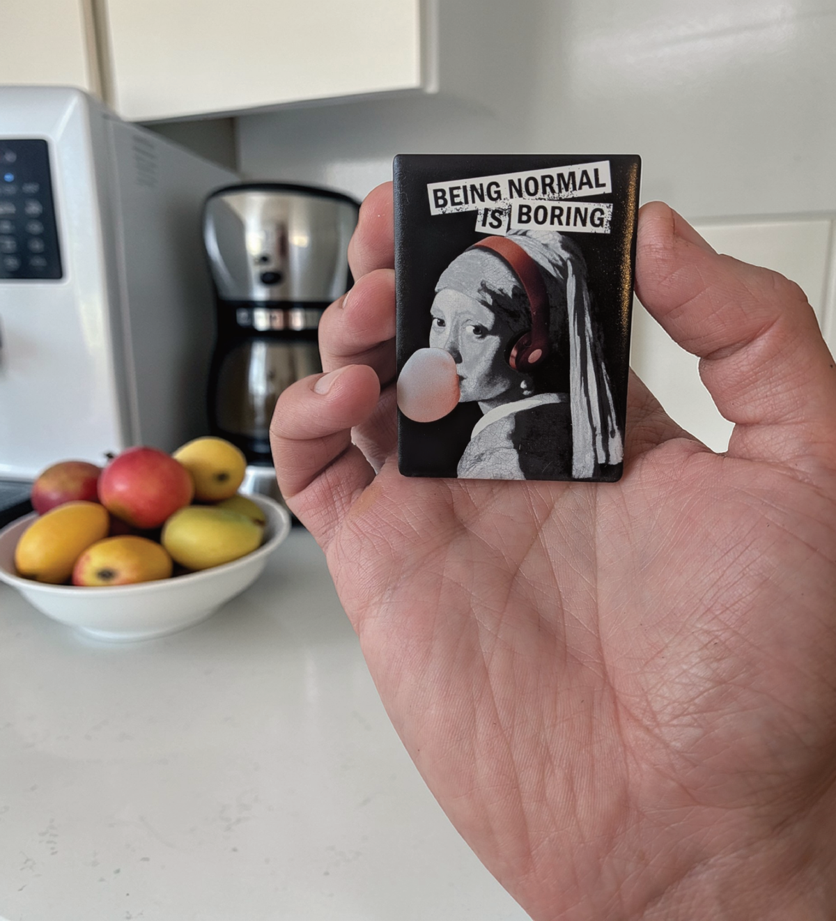 Rechteckiger Keramikmagnet (5x7 cm) mit schwarzem Hintergrund. Gezeigt wird das "Mädchen mit dem Perlenohrring" in Schwarz-Weiß, das rote Kopfhörer trägt und eine rosa Kaugummiblase macht. Darüber steht in weißer Schreibmaschinen-Schrift auf schwarzen Balken: "BEING NORMAL IS BORING".