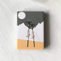 Rechteckiger Keramik-Kühlschrankmagnet (7x5 cm) mit zwei schwarzen Fahrrädern im Vordergrund vor weißen Bergen, einem graugrünen Himmel mit weißem Vollmond und einem ockergelben Boden mit spürbarem 3D-Relief.