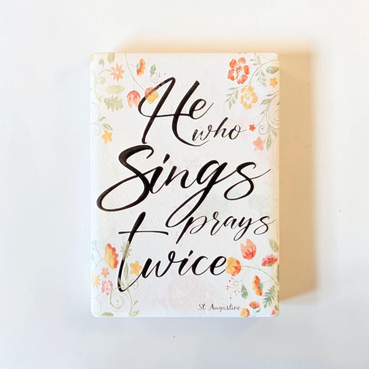 Rechteckiger Keramik-Kühlschrankmagnet (7x5 cm) mit seidenmatter Glasur. Auf hellem Grund mit zarten orange-gelben Aquarell-Blumen steht der schwarze Handlettering-Schriftzug 'Who sings prays twice' im spürbaren 3D-Relief