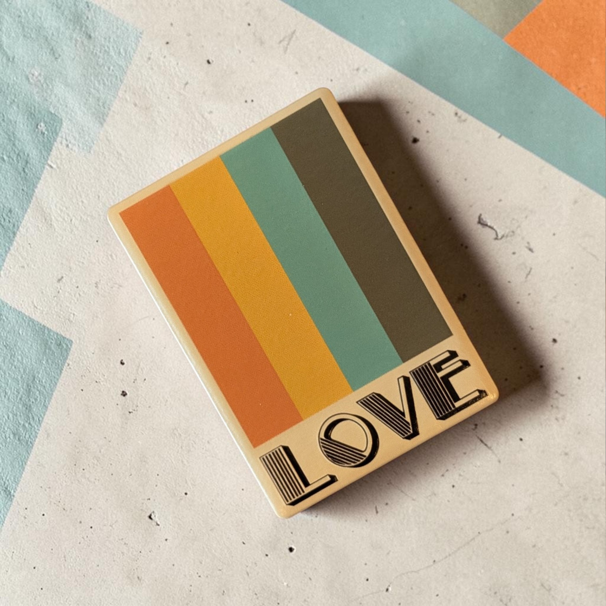 Rechteckiger Keramik-Kühlschrankmagnet (7x5 cm) mit seidenmatter Glasur. Auf beigem Grund verlaufen vier vertikale Streifen in Orange, Gelb, Petrol und Braun über dem schwarzen Schriftzug 'LOVE' im fühlbaren 3D-Relief.