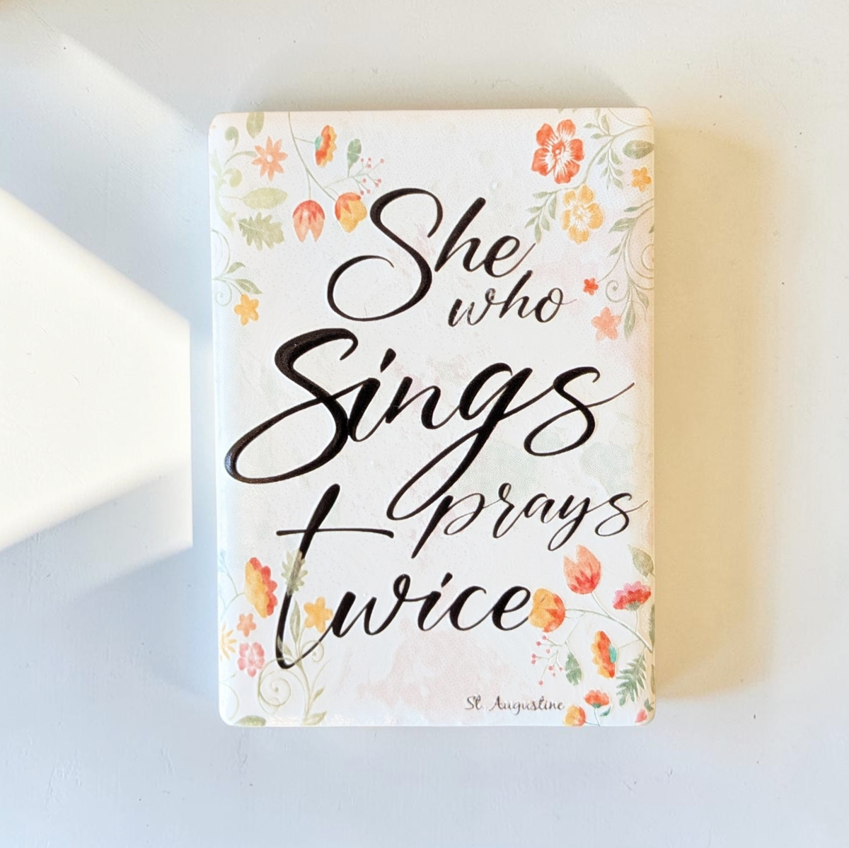 Rechteckiger Keramik-Kühlschrankmagnet (7x5 cm) mit seidenmatter Glasur. Auf hellem Grund mit zarten orange-gelben Aquarell-Blumen steht der schwarze Handlettering-Schriftzug 'Who sings prays twice' im spürbaren 3D-Relief