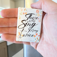 Rechteckiger Keramik-Kühlschrankmagnet (7x5 cm) mit seidenmatter Glasur. Auf hellem Grund mit zarten orange-gelben Aquarell-Blumen steht der schwarze Handlettering-Schriftzug 'Who sings prays twice' im spürbaren 3D-Relief