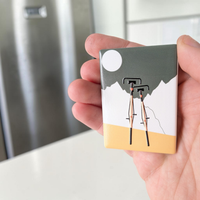 Rechteckiger Keramik-Kühlschrankmagnet (7x5 cm) mit zwei schwarzen Fahrrädern im Vordergrund vor weißen Bergen, einem graugrünen Himmel mit weißem Vollmond und einem ockergelben Boden mit spürbarem 3D-Relief.