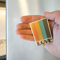 Rechteckiger Keramik-Kühlschrankmagnet (7x5 cm) mit seidenmatter Glasur. Auf beigem Grund verlaufen vier vertikale Streifen in Orange, Gelb, Petrol und Braun über dem schwarzen Schriftzug 'LOVE' im fühlbaren 3D-Relief.