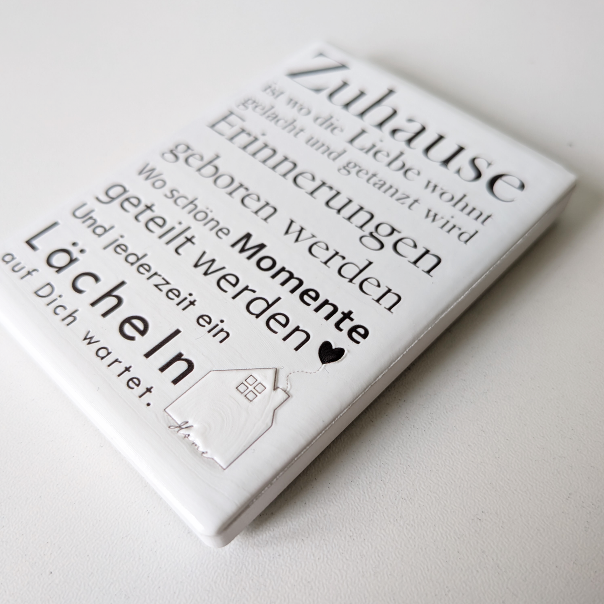 Rechteckiger Keramik-Kühlschrankmagnet (7x5 cm) mit seidenmatter Glasur. Auf hellem Grund steht der schwarze Spruch 'Zuhause ist wo die Liebe wohnt...' neben einer kleinen Haus-Illustration mit Herz im spürbaren 3D-Relief.