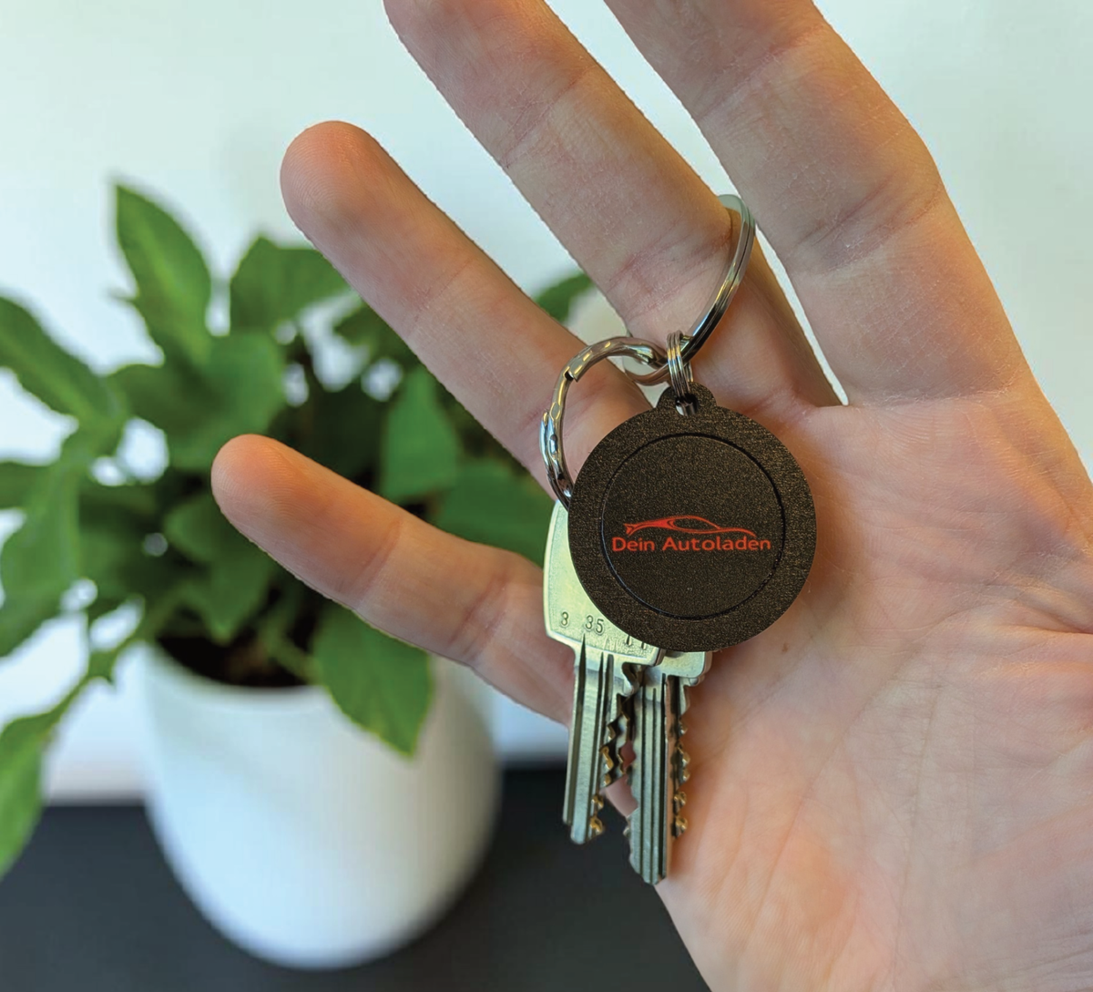 Hand hält einen runden, schwarzen NFC-Schlüsselanhänger aus Holz mit der
Aufschrift "DEIN LOGO -oder- NAME". Im Hintergrund eine kleine grüne Pflanze
auf hellem Tisch.