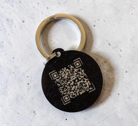 Hand hält einen runden, schwarzen NFC-Schlüsselanhänger aus Holz mit der
Aufschrift "DEIN LOGO -oder- NAME". Im Hintergrund eine kleine grüne Pflanze
auf hellem Tisch.