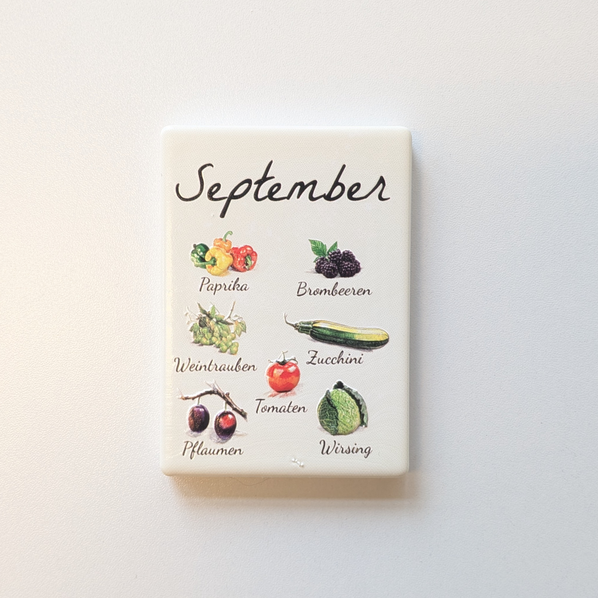 Saisonkalender Kühlschrankmagneten aus Keramik. Jeder Magnet repräsentiert einen Monat von Januar bis Dezember mit deutscher Beschriftung in geschwungener Schrift. Auf der cremefarbenen Oberfläche sind detailreiche, farbige Illustrationen von saisonalem Obst und Gemüse mit den jeweiligen Namen abgebildet.