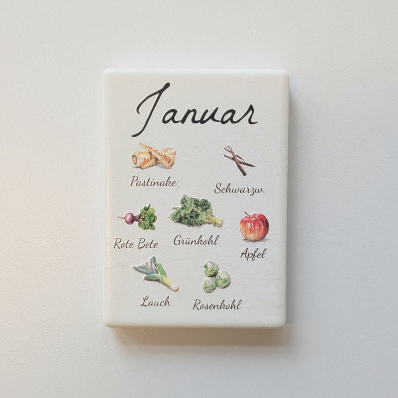 Set aus 12 rechteckigen Kühlschrankmagneten aus Keramik, die ordentlich in Reihen nebeneinander angeordnet sind. Jeder Magnet repräsentiert einen Monat von Januar bis Dezember mit deutscher Beschriftung in geschwungener Schrift. Auf der cremefarbenen Oberfläche sind detailreiche, farbige Illustrationen von saisonalem Obst und Gemüse mit den jeweiligen Namen abgebildet.